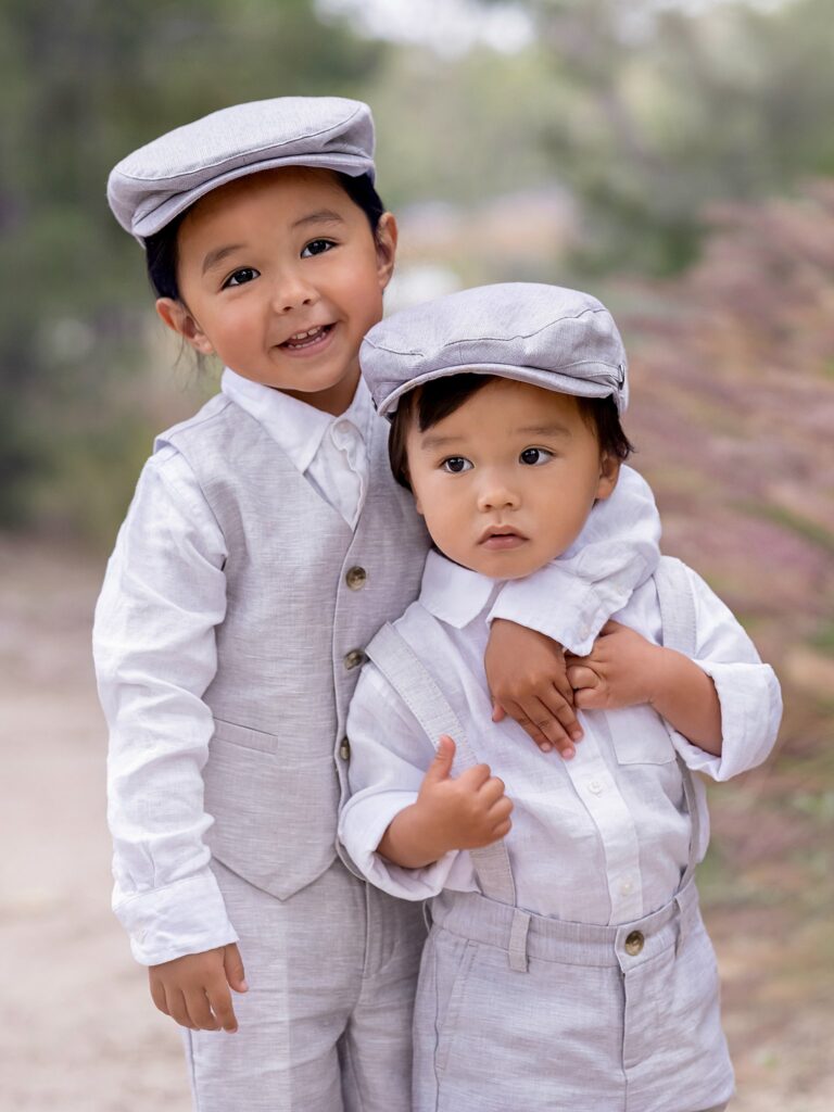 Two toddler brothers stand in a park path in matching white and grey hats and outfits
