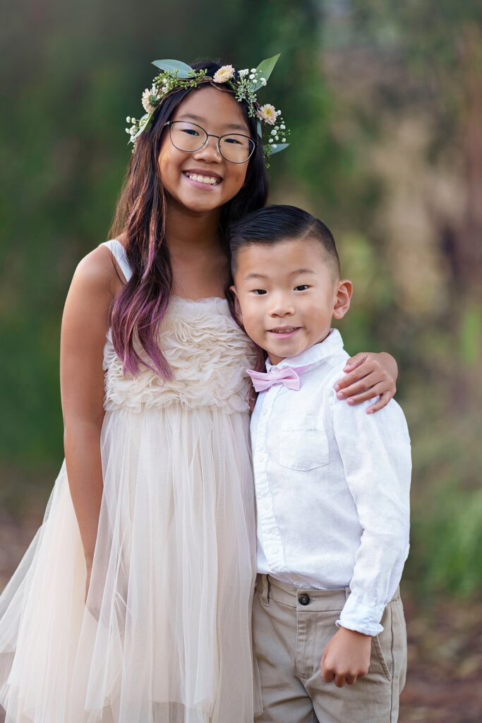 A young girl in a white dress and floral headband stands with her arm around her younger brother in a pink bowtie and white shirt after attending Best private schools in Orange County