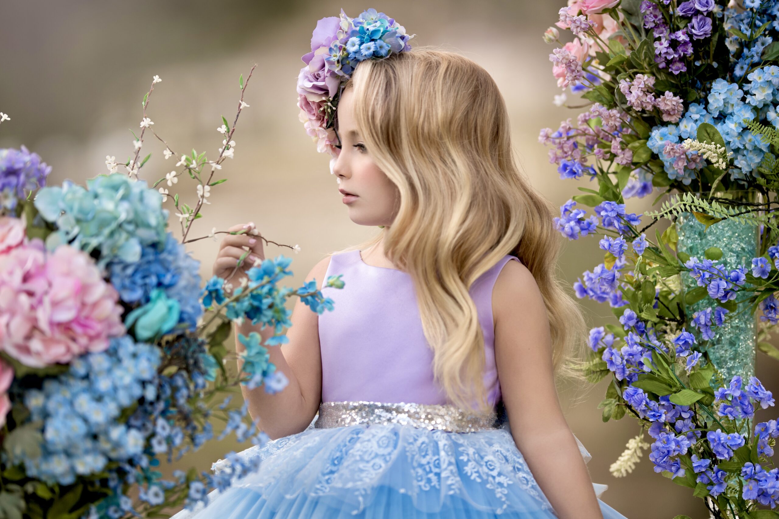 Girl in purple and light blue dress looking at purple and blue flowers with a purple and blue floral headpiece on her head.