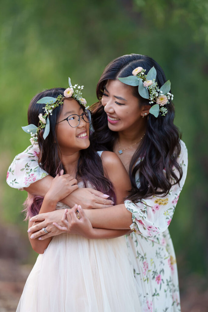 Asian mom embracing her 8-year-old daughter from behind while they are both wearing floral crowns with eucalyptus leaves and vintage pom flowers and are standing in front of some greenery outdoors. 