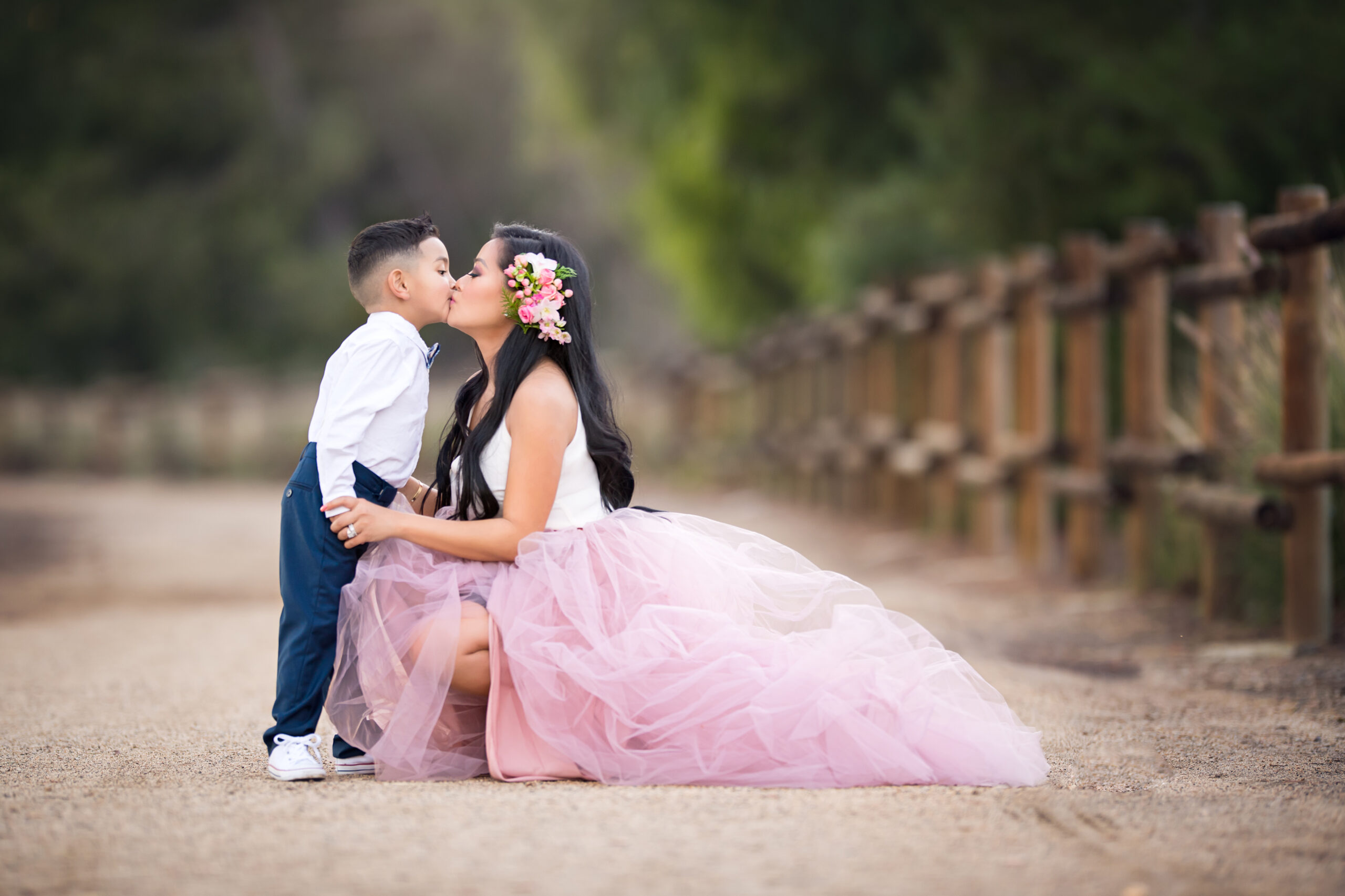 Woman with long black hair with a floral accessory on the side in pink tutu and white tank top kneeling on a dirt path and kissing a young boy in a white button up shirt and navy blue pants.
