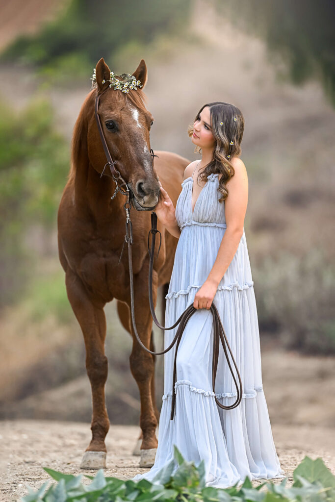 Teenage girl in light blue dress touching her brown horse that is standing beside her on the muzzle while holding his reins in her hands and staring at him lovingly. 