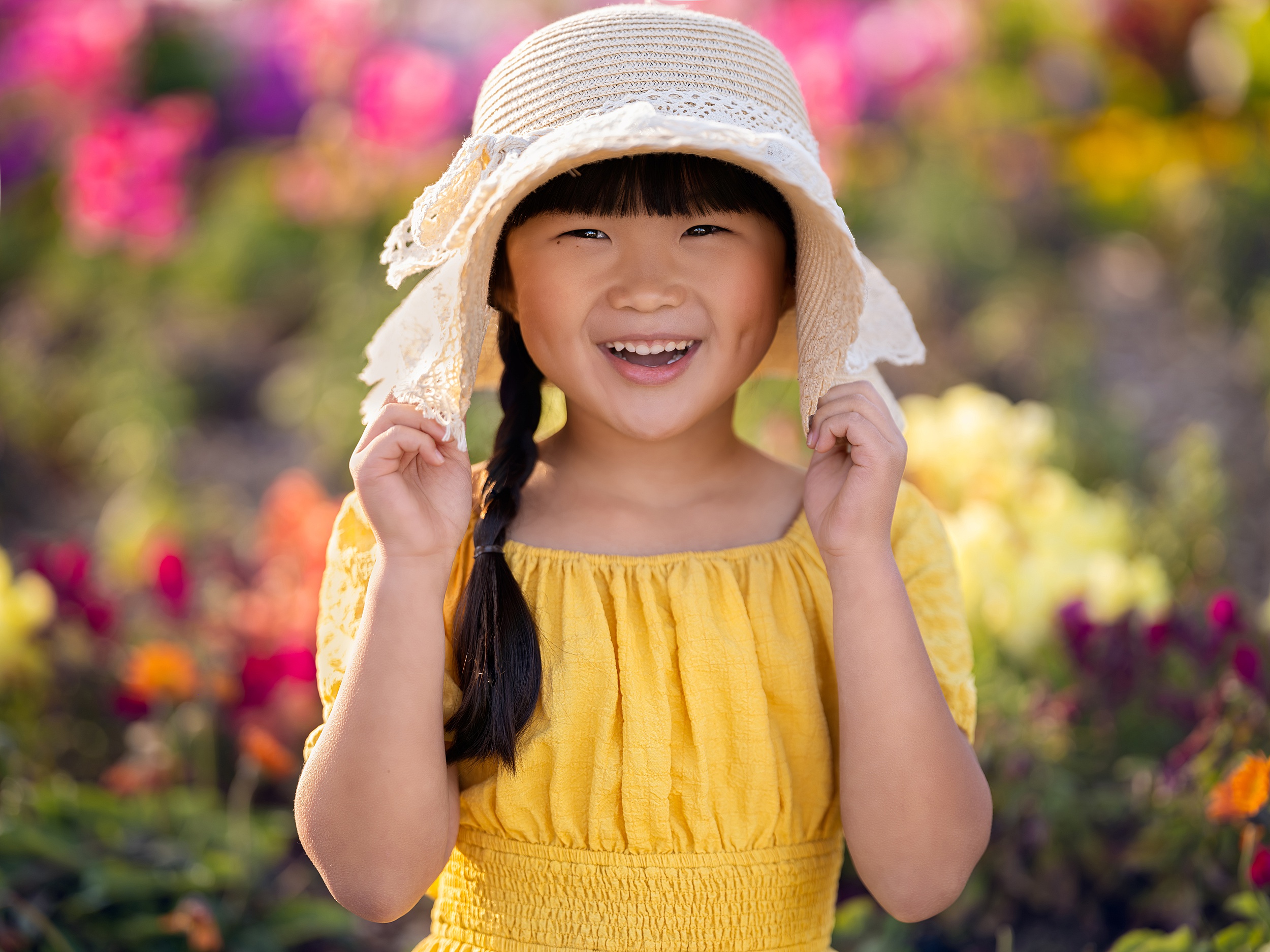 A young girl in a yellow dress pulls her woven hat over her ears while standing in a flower garden