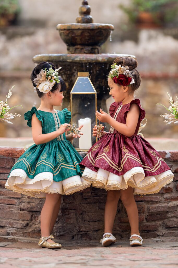 Two young girl stand by a fountain in matching red and green dresses and hair pieces after visiting orange county baby boutiques
