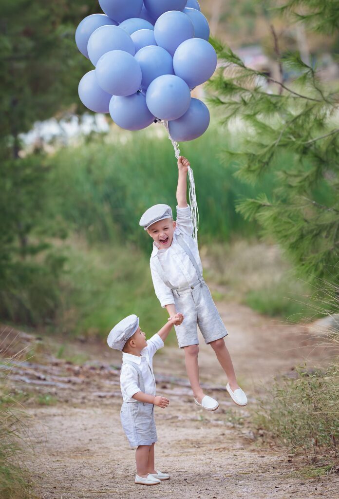 A young boy in suspenders is lifted off the ground by a bundle of balloons while holding hands with little brother thanks to orange county baby boutiques